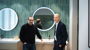 Δύο άνδρες σε σύγχρονο μπάνιο showroom, μπροστά από στρογγυλούς φωτιζόμενους καθρέφτες και πράσινη κεραμική επένδυση τοίχου.